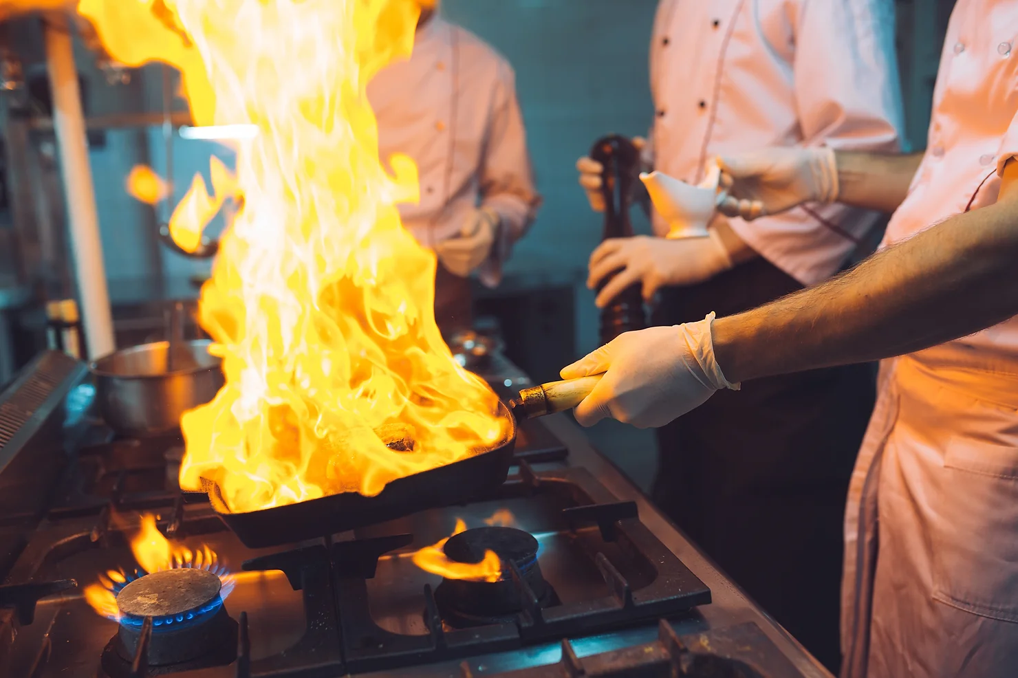 A person is cooking with a large flame bursting from a pan on a gas stove, with other people and kitchen equipment in the background.