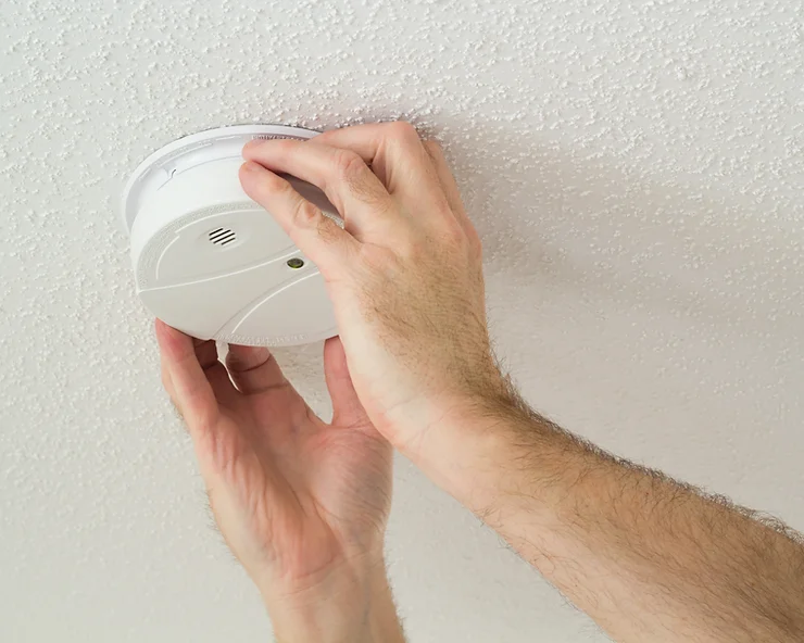 A person is installing a white smoke detector on a textured ceiling, using both hands to secure the device in place.
